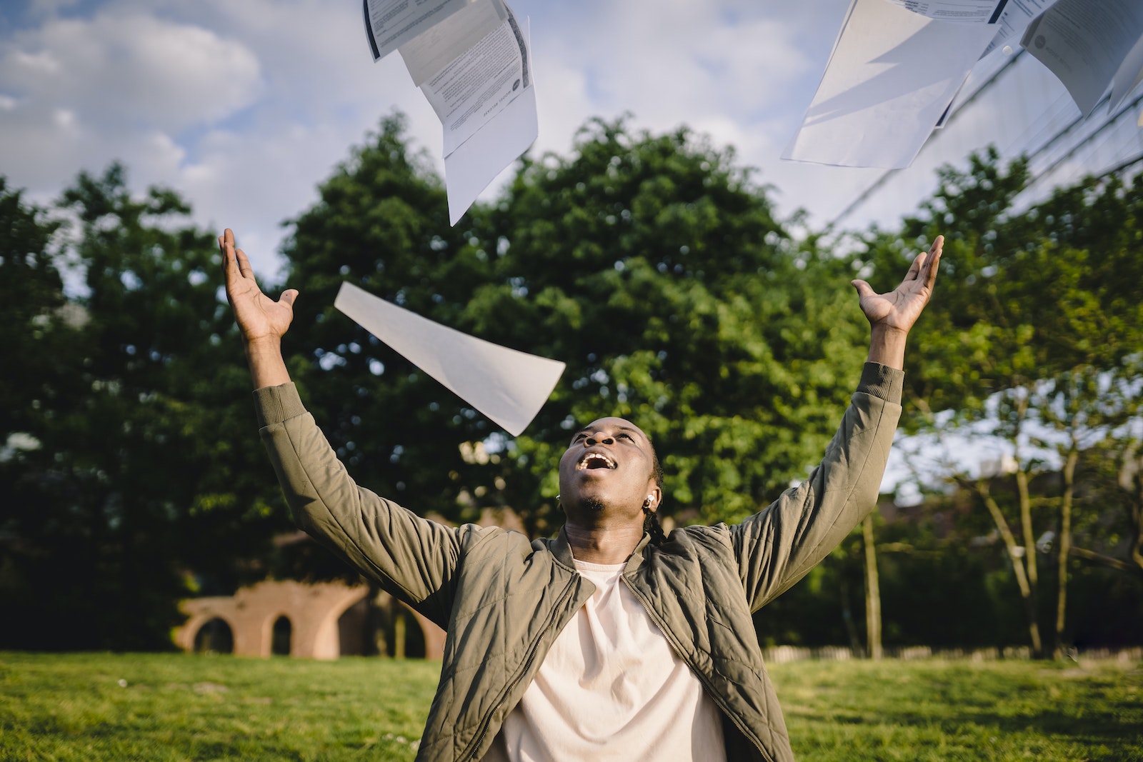 Cheerful young African American male student in casual clothes throwing college papers up in air while having fun in green park after end of exams